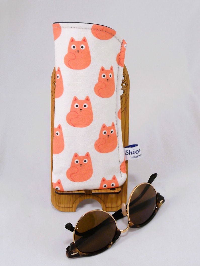 Puede incluir: Estuche para gafas de sol de tela con estampado de gatos blancos y naranjas con ribete negro. El estuche est&aacute; sobre un soporte de madera. Un par de gafas de sol de carey marr&oacute;n est&aacute;n sobre una superficie blanca.