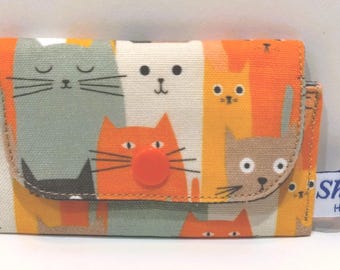 May include: A small, orange and white cat-themed wallet with a snap closure. The wallet features a repeating pattern of cartoon cats in various colors. A small metal ring is attached to the side. The tag reads "Shio Handmade".