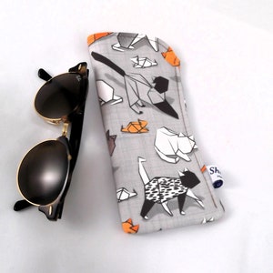 Puede incluir: Un estuche para gafas de tela gris con un patr&oacute;n geom&eacute;trico de gatos origami negros y naranjas. El estuche tiene una etiqueta blanca con el texto "Shop Handmade".