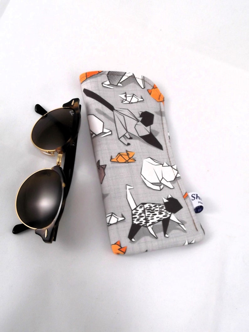 Puede incluir: Un estuche para gafas de tela gris con un patr&oacute;n geom&eacute;trico de gatos origami negros y naranjas. El estuche tiene una etiqueta blanca con el texto "Shop Handmade".