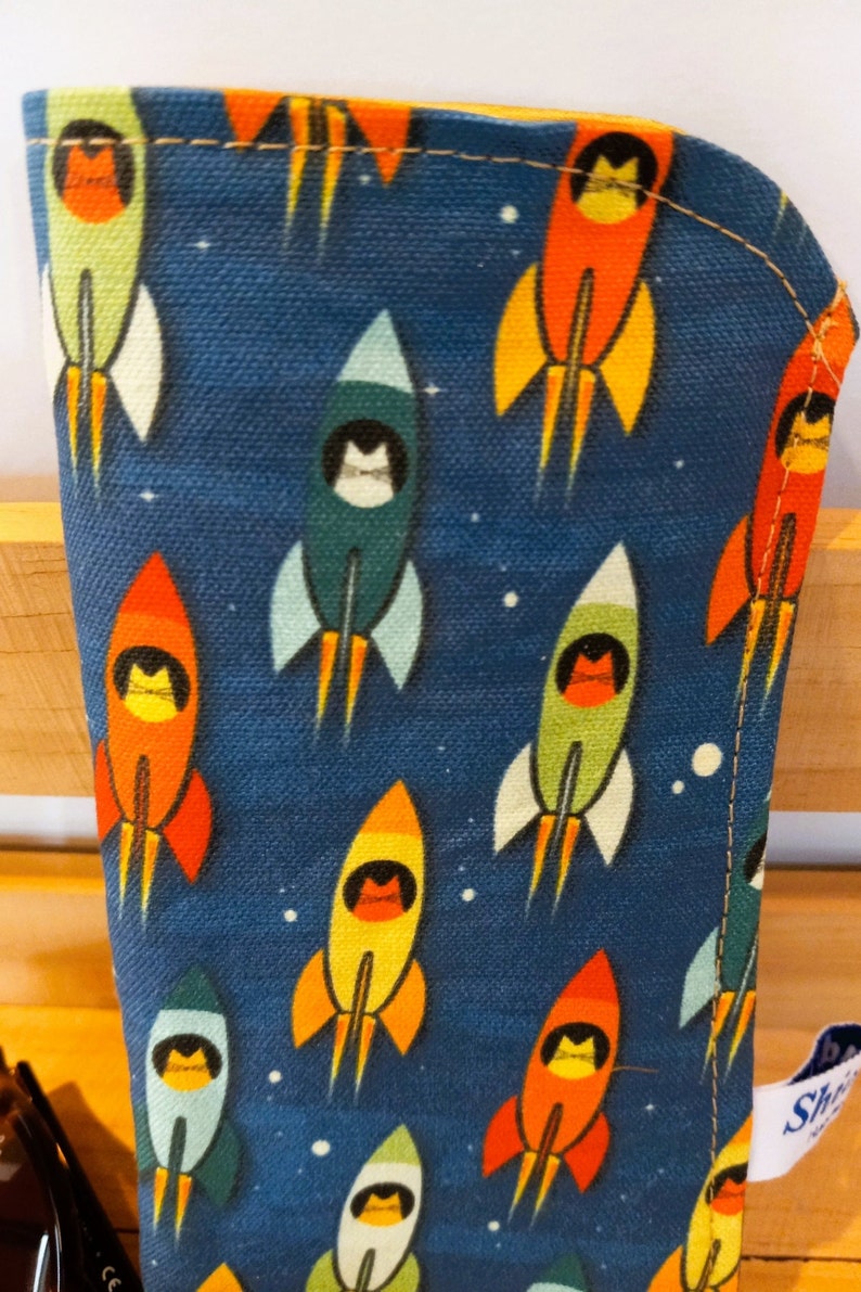 Puede incluir: Una bolsa de lona azul con cremallera con un patr&oacute;n repetido de cohetes con gatos en las ventanas. Los cohetes son rojos, naranjas, amarillos, verdes y azules. El fondo es azul oscuro con estrellas blancas.