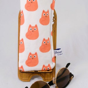 Puede incluir: Un estuche para gafas de estampado de gatos blanco y naranja con una etiqueta azul que dice "Shio! Hecho a mano". El estuche est&aacute; sobre un soporte de madera con unas gafas de sol de carey marr&oacute;n delante.