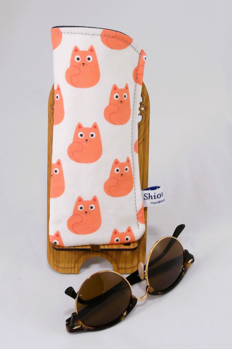 Puede incluir: Un estuche para gafas de estampado de gatos blanco y naranja con una etiqueta azul que dice "Shio! Hecho a mano". El estuche est&aacute; sobre un soporte de madera con unas gafas de sol de carey marr&oacute;n delante.