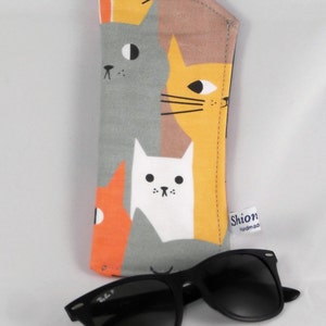 Puede incluir: Un estuche para gafas de tela gris, naranja, amarillo y blanco con un patr&oacute;n de gatos. El estuche tiene una cremallera negra y una peque&ntilde;a etiqueta que dice "Shion handmade". Un par de gafas de sol negras est&aacute;n junto al estuche.
