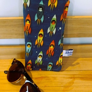 Puede incluir: Un estuche para gafas de tela azul y amarilla con un patr&oacute;n de cohetes. El estuche tiene una cremallera amarilla y una etiqueta blanca que dice "Shio Handmade".