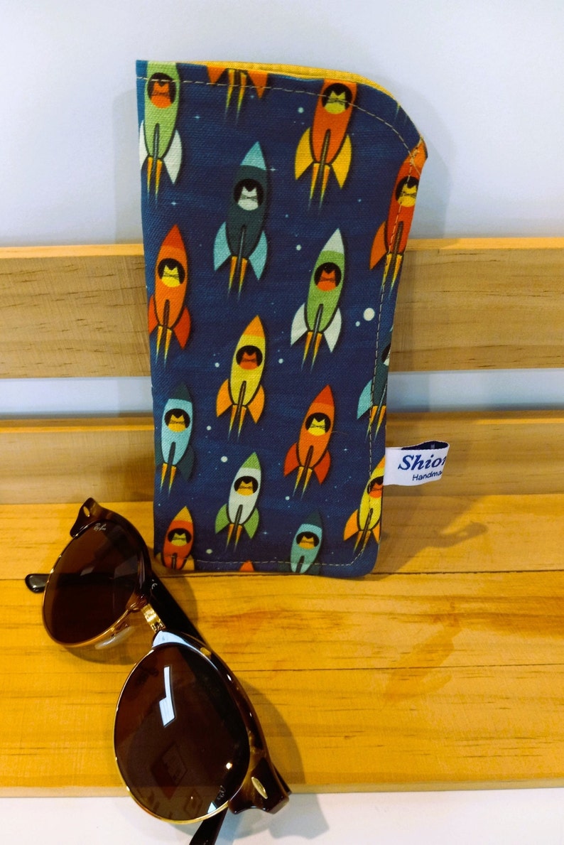 Puede incluir: Un estuche para gafas de tela azul y amarilla con un patr&oacute;n de cohetes. El estuche tiene una cremallera amarilla y una etiqueta blanca que dice "Shio Handmade".