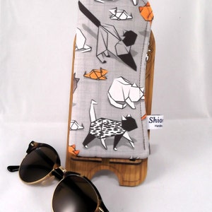 Puede incluir: Un estuche para gafas de tela gris con un patr&oacute;n de gatos origami en negro, blanco y naranja. El estuche est&aacute; en un soporte de madera. El texto "Shiori Handmade" est&aacute; en una etiqueta blanca unida al estuche.