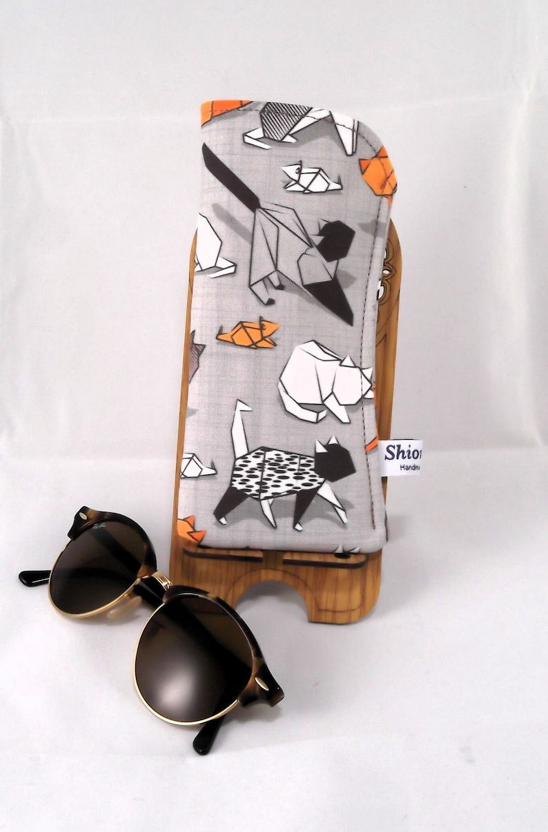 Puede incluir: Un estuche para gafas de tela gris con un patr&oacute;n de gatos origami en negro, blanco y naranja. El estuche est&aacute; en un soporte de madera. El texto "Shiori Handmade" est&aacute; en una etiqueta blanca unida al estuche.