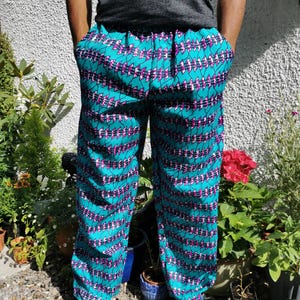 Op de afbeelding: Turquoise broek met een geometrisch patroon in roze en witte tinten. De broek heeft een elastische tailleband en is gemaakt van een lichtgewicht stof.