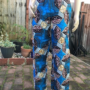 Può includere: Pantaloni blu fantasia con un design geometrico. I pantaloni presentano un motivo vibrante con sfumature di blu, marrone e bianco. I pantaloni sono esposti su un manichino all'aperto.