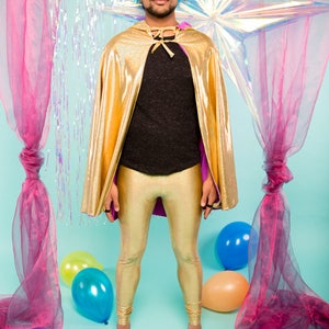 May include: A person wearing a gold cape and gold leggings stands in front of a blue background with pink and iridescent curtains and balloons.