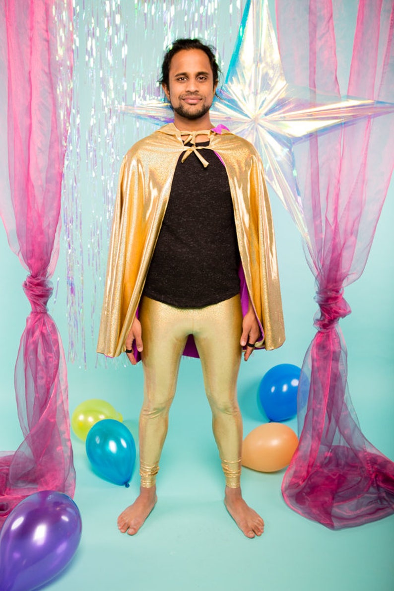 May include: A person wearing a gold cape and gold leggings stands in front of a blue background with pink and iridescent curtains and balloons.