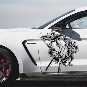 May include: A white car with a black and white decal of a demon skull, snake, and swords on the side.