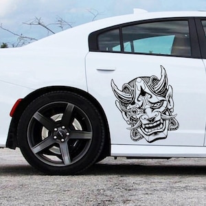 May include: A white car with a black and white sticker of a demon mask on the side. The sticker is detailed and has a mechanical look.