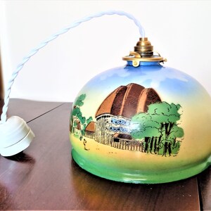 Peut inclure: Un luminaire suspendu en céramique vintage avec une scène peinte en bleu, vert et marron d'un bâtiment avec un toit marron et des arbres. Le luminaire a un cordon blanc et une douille en céramique blanche.