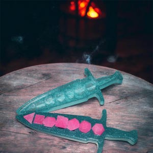 Puede incluir: Dos soportes para dados con forma de daga de resina brillante de color verde azulado. Uno contiene dados transparentes, el otro dados rosas. Las dagas están sobre una superficie de madera, con un fondo borroso de chimenea.