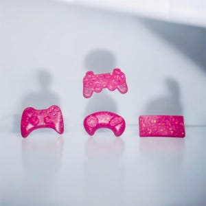 May include: Four pink, glittery game controller-shaped items. There are two traditional controllers, one with a rounded top, and a rectangular item. The items are set against a white background.