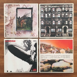 May include: Set of four square ceramic coasters featuring album covers from Led Zeppelin. The coasters feature images of a house, a blimp, a wall with a picture frame, and a rocky landscape.