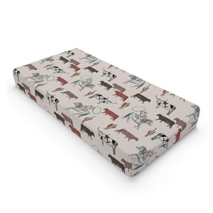 May include: A rectangular, beige baby changing pad with a repeating pattern of cowboys, bulls, cacti, and speech bubbles in brown, black, and turquoise. The pad is designed for infant care.
