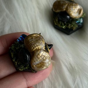 May include: A handcrafted miniature sculpture featuring two gold-colored acorns with textured surfaces, set on a dark base with moss and a black stone. The sculpture is held in a hand against a white, fluffy background.