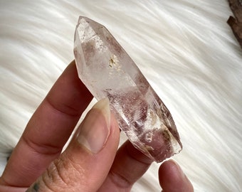 May include: A clear quartz crystal with a pink hue and inclusions. The crystal is pointed at both ends and has a smooth, polished surface.
