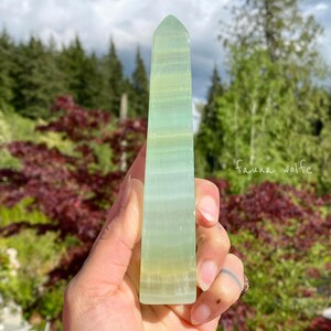 Puede incluir: Un cristal en forma de obelisco verde y blanco con una superficie lisa y pulida. El cristal se sostiene en una mano.