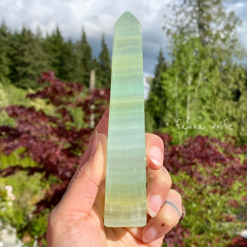 Puede incluir: Un cristal en forma de obelisco verde y blanco con una superficie lisa y pulida. El cristal se sostiene en una mano.