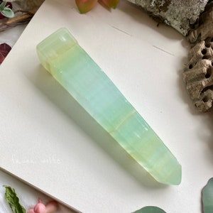 Puede incluir: Una punta de cristal verde claro y blanco con una superficie lisa y pulida. El cristal tiene la forma de una pir&aacute;mide larga y delgada.