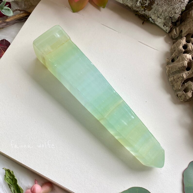 Puede incluir: Una punta de cristal verde claro y blanco con una superficie lisa y pulida. El cristal tiene la forma de una pir&aacute;mide larga y delgada.