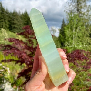 Puede incluir: Una punta de cristal verde claro y blanco, tambi&eacute;n conocida como varita, se sostiene en una mano. El cristal es liso y pulido.