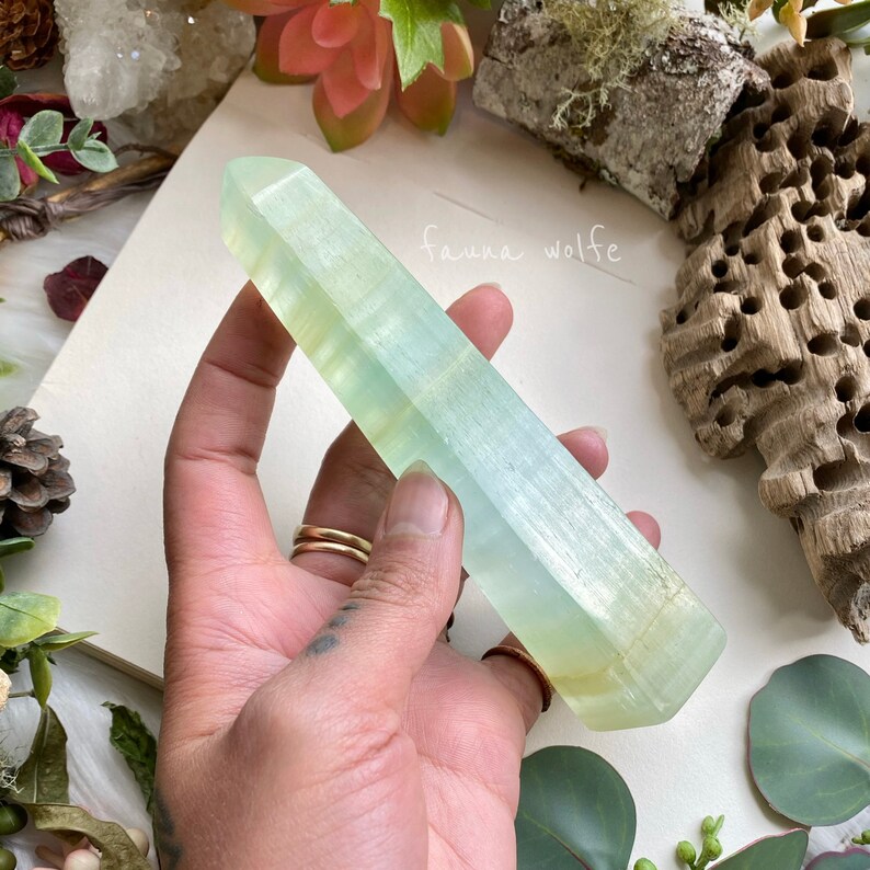 Puede incluir: Una punta de cristal transl&uacute;cida verde claro con una superficie lisa y pulida. El cristal mide aproximadamente 15 cm de largo y tiene una punta puntiaguda. El cristal se sostiene en una mano, con el fondo desenfocado.