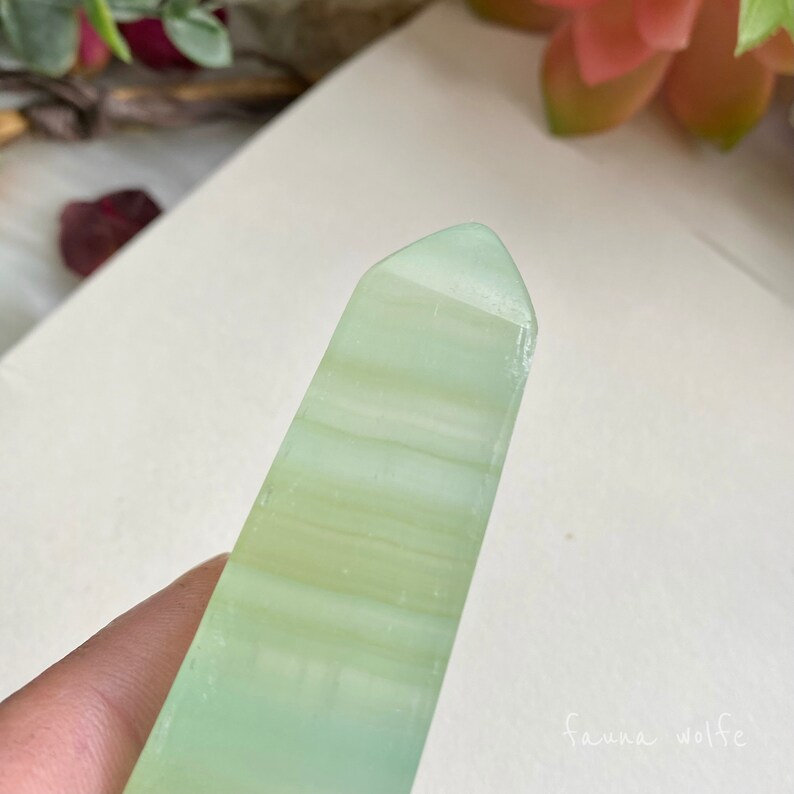 Puede incluir: Un cristal puntiagudo, transl&uacute;cido, pulido y de color verde claro con rayas blancas. El cristal se sostiene en una mano.