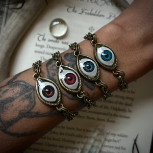 May include: Four eye-shaped charms on a bronze chain bracelet. The charms are each a different color: red, blue, teal, and brown. The bracelet is worn on a person's wrist.