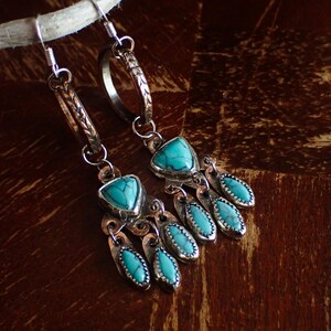 May include: A pair of silver-toned earrings with turquoise-colored stones. The earrings feature a triangular stone at the top and several teardrop-shaped stones hanging below. The earrings are displayed on a wooden surface.