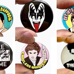 May include: Six circular pin badges with pop culture references. The badges feature images of musicians and phrases including "Gay as a Daffodil" and "Take on Me".