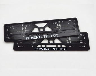 Puede incluir: Dos marcos negros para matrículas con la inscripción "PERSONALIZED TEXT" en blanco. Los marcos son rectangulares con un diseño geométrico y están apilados uno encima del otro.