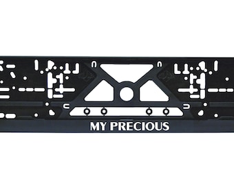 Puede incluir: Marco negro para matrícula con el texto blanco "MY PRECIOUS". El marco tiene forma rectangular con varios recortes y una zona de montaje central. Diseñado para una placa de matrícula de tamaño estándar.
