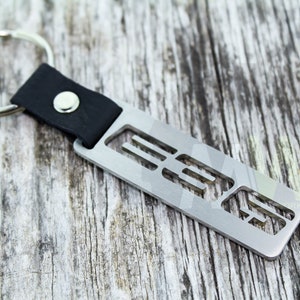 May include: A silver metal keychain with a black leather strap and a silver ring. The metal part of the keychain has a cut-out design of the letters "E30".
