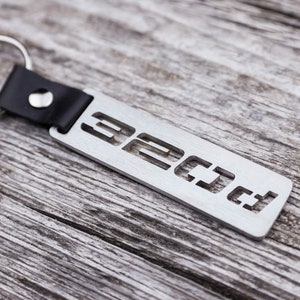 Op de afbeelding: Een zilveren metalen sleutelhanger met een zwart leren bandje en de tekst "32 CJ 4" uitgesneden.