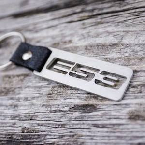 Puede incluir: Un llavero de metal plateado con una correa de cuero negro y la palabra "E53" recortada en el metal.