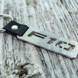 Puede incluir: Un llavero de metal plateado con una correa de cuero negro y un diseño recortado de las letras "FTG".