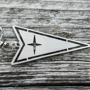 May include: A silver metal keychain with a star-shaped design in the center of a triangular shape. The keychain has a silver metal ring.