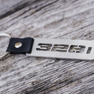 May include: A silver metal keychain with the cut-out text "328i" attached to a black leather strap and a silver key ring. The keychain is displayed on a weathered wooden surface.