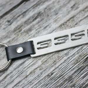 Könnte beinhalten: Ein silberner Schlüsselanhänger aus Metall mit einem schwarzen Lederband und einem Metallring. Das Metallschild hat die Nummer "335i" ausgeschnitten.