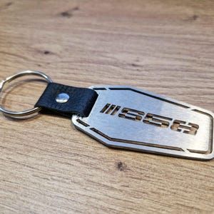 Può includere: Un portachiavi in metallo argentato con le lettere "SSE3" ritagliate. Il portachiavi ha un cinturino in pelle nera e un portachiavi argentato. Lo sfondo è una superficie in legno.