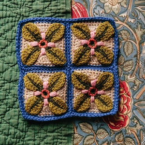 May include: Four crocheted squares with a floral design. Each square features a pink flower with a green center surrounded by four green leaves. The squares are joined together with blue yarn.