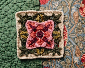 May include: A square, handmade crochet piece featuring a central flower design in shades of pink and red, surrounded by green leaves and a beige border. The piece is set against a green quilted background and a floral patterned fabric.