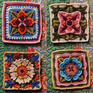 May include: Four crocheted squares with floral designs in shades of pink, red, blue, and green. The squares are arranged in a grid pattern on a fabric background.