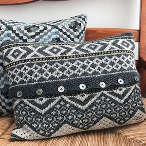 Puede incluir: Dos almohadas decorativas con un patrón geométrico en tonos grises, blancos y negros. Las almohadas tienen una textura de punto y están acentuadas con botones.