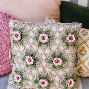 Op de afbeelding: Een gehaakt kussen met een beige achtergrond en bloemmotieven. Het kussen heeft een herhalend patroon van bloemen met gehaakte details in roze, paars en groen.